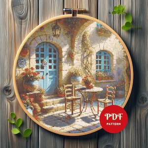 May include: A cross-stitch pattern of a European courtyard scene, featuring a blue door, windows, and a table with chairs. The scene is framed in a wooden embroidery hoop. Red flowers in pots and climbing vines add color. The text "PDF PATTERN" is in a red circle.