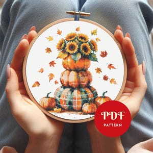 Op de afbeelding: Kruissteekborduurwerk in een houten hoepel met een stapel pompoenen, bekroond met zonnebloemen en herfstbladeren. De pompoenen zijn oranje en groen, met een ruitpatroon op de onderste pompoen. Extra kleine pompoenen en bladeren zijn verspreid.