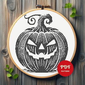 May include: A black and white cross-stitch design of a carved pumpkin face, presented in a wooden embroidery hoop. The pumpkin features a sinister grin and triangular eyes. A red circle with "PDF PATTERN" is in the lower right.