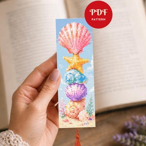 May include: A cross-stitch bookmark featuring a stack of seashells and a starfish in shades of pink, yellow, blue, and purple. The bookmark has a tassel and the text "PDF PATTERN" in a red circle.
