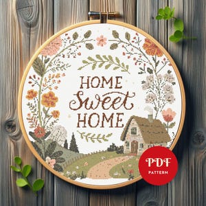 Puede incluir: Un bordado de punto de cruz en un bastidor de madera redondo con el texto HOME SWEET HOME en letras marrones. El diseño incluye una corona floral con elementos botánicos naranjas, rosas y verdes y una pequeña casa con un camino sobre tela blanca.