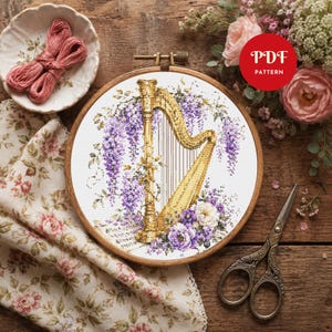 Könnte beinhalten: Ein Kreuzstichmuster in einem hölzernen Stickrahmen mit einer goldenen Harfe, umgeben von violettem Blauregen und weißen Blumen. Ein rotes kreisförmiges Abzeichen mit dem weißen Text PDF PATTERN ist oben rechts im Bild zu sehen.