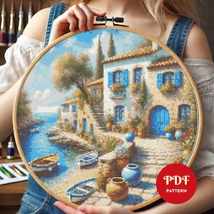 May include: A completed cross-stitch project in a wooden hoop depicts a coastal scene with buildings, boats, and water. The buildings have blue shutters and doors. The scene is in shades of blue, beige, and brown. A red circle with the text "PDF PATTERN" is in the lower right.