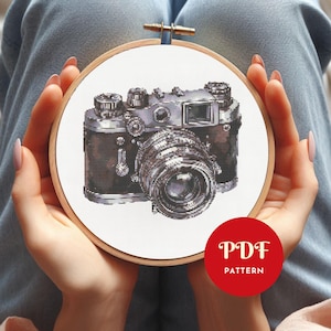 Puede incluir: Un bordado de punto de cruz de una cámara vintage en un aro de madera. La cámara está detallada con tonos plateados y marrones. Un círculo rojo con el texto "PDF PATTERN" está en la esquina inferior derecha.