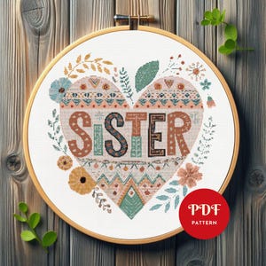 May include: A cross-stitch design in a wooden hoop displays the word "SISTER" within a heart shape. The heart is adorned with floral and geometric patterns in orange, teal, and brown hues. A red circle with "PDF PATTERN" is in the lower right.