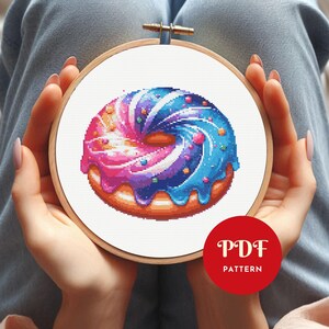 May include: A cross-stitch pattern of a colorful donut with blue, pink, and purple frosting and sprinkles. The donut is set in a wooden embroidery hoop. A red circle with the text "PDF PATTERN" is in the lower right corner.