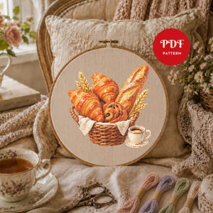 K&ouml;nnte beinhalten: Ein Kreuzstich-Stickmuster in einem Holzrahmen, das einen geflochtenen Korb voller goldbrauner Croissants, ein Baguette und Weizen&auml;hren sowie eine kleine Tasse Kaffee zeigt. Ein rotes kreisf&ouml;rmiges Abzeichen enth&auml;lt den Text PDF PATTERN.