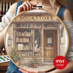 May include: A cross-stitch pattern of a bookstore, featuring the word "BOOKSTORE" above the shop's facade. The design includes shelves of books, potted plants, and a detailed door. The pattern is held in a wooden embroidery hoop.