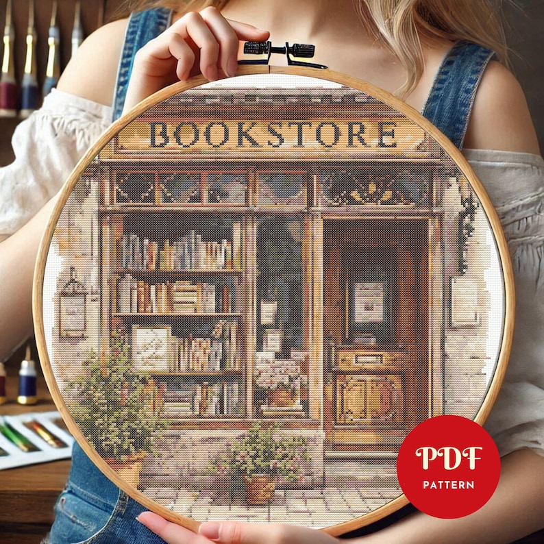 May include: A cross-stitch pattern of a bookstore, featuring the word "BOOKSTORE" above the shop's facade. The design includes shelves of books, potted plants, and a detailed door. The pattern is held in a wooden embroidery hoop.