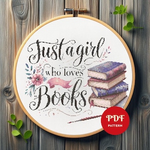 Pode incluir: Bordado em ponto cruz em um bastidor de madeira com o texto "Just a girl who loves Books". O design inclui uma pilha de livros, detalhes florais e uma fita rosa. Um círculo vermelho com "PDF PATTERN" está no canto inferior direito.