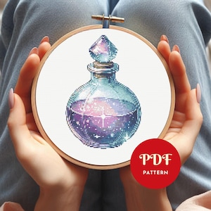 May include: A cross-stitch pattern of a potion bottle with a diamond-shaped stopper. The bottle is filled with a galaxy-like liquid in shades of purple, pink, and blue. The design is set in a wooden embroidery hoop. A red circle with the text "PDF PATTERN" is in the lower right.
