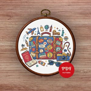 May include: A cross-stitch design in a wooden hoop depicts a blue and red suitcase adorned with travel stickers, a camera, passport, sunglasses, and travel icons. The text "PDF PATTERN" is in a red circle.
