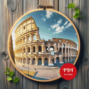 Pode incluir: Bordado em ponto cruz do Coliseu em Roma, Itália, num bastidor de madeira. O design detalhado apresenta o icónico anfiteatro sob um céu azul com nuvens. Um círculo vermelho com "PDF PATTERN" está no canto inferior direito.