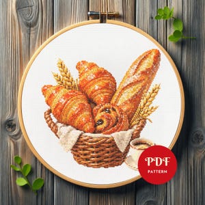 K&ouml;nnte beinhalten: Ein Kreuzstich-Stickprojekt in einem Holzrahmen, das einen geflochtenen Korb voller goldbrauner Croissants, ein Baguette und Weizen&auml;hren zeigt. Eine kleine Kaffeetasse steht neben dem Korb. Ein rotes rundes Abzeichen zeigt den Text PDF PATTERN.