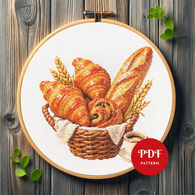 K&ouml;nnte beinhalten: Ein Kreuzstich-Stickprojekt in einem Holzrahmen, das einen geflochtenen Korb voller goldbrauner Croissants, ein Baguette und Weizen&auml;hren zeigt. Eine kleine Kaffeetasse steht neben dem Korb. Ein rotes rundes Abzeichen zeigt den Text PDF PATTERN.