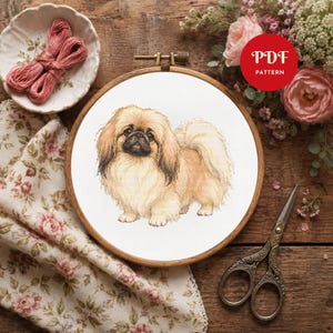 May include: A cross stitch pattern featuring a tan and cream Pekingese dog centered in a wooden embroidery hoop. The design includes a red circular badge with the white text PDF PATTERN, pink embroidery floss, floral fabric, and metal scissors.