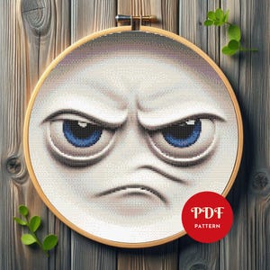 May include: A cross-stitch pattern of an angry face with blue eyes, set in a wooden embroidery hoop. The design features a frowning mouth and furrowed brow. A red circle with the text "PDF PATTERN" is in the lower right corner.