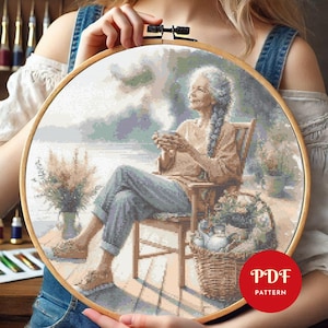 May include: A cross stitch pattern depicting a woman seated in a chair on a veranda, holding a cup of tea. She gazes out at a lake with mountains in the background. The pattern is displayed in a wooden embroidery hoop.