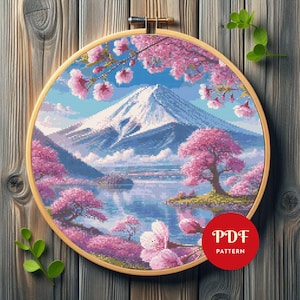 May include: A cross-stitch pattern depicting a Japanese landscape with Mount Fuji, cherry blossoms, and a lake, set within a wooden embroidery hoop. The design uses pink, blue, and white hues. A red circle with "PDF PATTERN" is in the lower right corner.