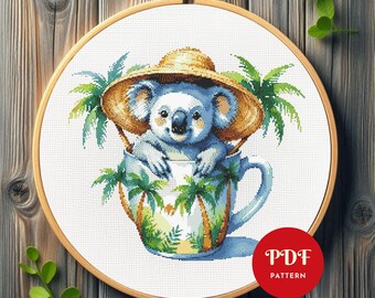 May include: Cross-stitch design of a koala wearing a straw hat, nestled in a teacup adorned with palm trees. The artwork is presented in a wooden embroidery hoop. A red circle with "PDF PATTERN" text is in the lower right corner.