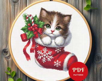 May include: Cross-stitch pattern featuring a kitten in a Christmas stocking. The red stocking has white snowflake designs. The kitten is brown and white, with green eyes, and is holding a sprig of holly and a candy cane. The pattern is in a wooden embroidery hoop.