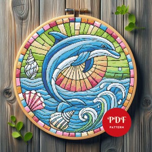 May include: A cross-stitch pattern of a dolphin leaping through waves, set within a colorful, stained-glass style design. The image includes seashells and a circular wooden embroidery hoop. The text "PDF PATTERN" is in a red circle.