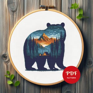 Peut inclure: Motif de point de croix représentant une silhouette d'ours avec un paysage de montagne et de forêt. Le design utilise des nuances de bleu, d'orange et de vert, dans un cercle à broder en bois. Un cercle rouge avec "PDF PATTERN" est visible.