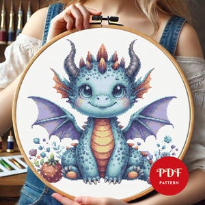 Könnte beinhalten: Kreuzstichmuster mit einem niedlichen blauen Drachen mit violetten Flügeln und gelbem Bauch. Der Drache sitzt auf einem Blumenbeet und hat einen freundlichen Ausdruck. Das Muster ist als PDF-Download erhältlich.