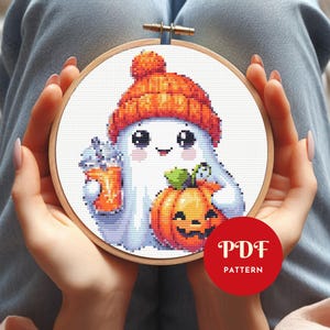 Könnte beinhalten: Kreuzstichmuster eines freundlichen Geistes mit orangefarbener Strickmütze, der einen Kürbis und ein Getränk hält. Das Design befindet sich in einem hölzernen Stickrahmen. Ein roter Kreis mit dem Text "PDF PATTERN" ist sichtbar.