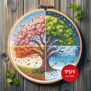 Puede incluir: Un bastidor de bordado de punto de cruz con un árbol dividido en cuatro secciones, cada una representando una estación: primavera con flores rosas, verano con hojas verdes, otoño con hojas naranjas e invierno con nieve. El texto "PDF PATTERN" está en un círculo rojo.