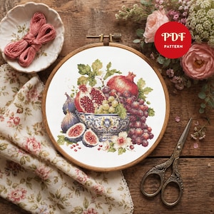 Peut inclure: Un modèle de point de croix dans un tambour à broder en bois représentant un bol rempli de grenades, de raisins et de figues. Le motif inclut des feuilles vertes et de petites fleurs blanches. Un badge rouge avec le texte PDF PATTERN est visible.