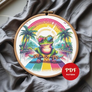 May include: Cross-stitch pattern in a wooden hoop featuring a frog wearing sunglasses and sitting on a skateboard. The design includes a rainbow, palm trees, and the text "PDF PATTERN" in a red circle.