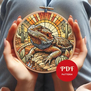 May include: A cross-stitch design of a bearded dragon in a desert scene, held in a wooden embroidery hoop. The image shows a detailed lizard in brown, orange, and blue hues, with cacti and a sunset in the background. The text "PDF PATTERN" is visible.