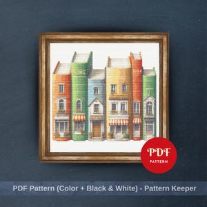 May include: A cross-stitch pattern featuring a row of colorful books designed to look like a street of buildings, with the words HOME and BOOK visible on the spines, framed in a wooden border with a red circular PDF PATTERN label.