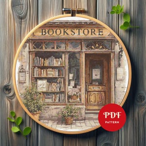 May include: A cross-stitch pattern of a bookstore, framed in a wooden hoop. The shop front features a detailed facade with the word "BOOKSTORE" above the window. Bookshelves are visible through the windows, and potted plants flank the entrance. A red circle with "PDF PATTERN" is in the lower right.