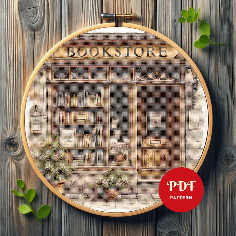 May include: A cross-stitch pattern of a bookstore, framed in a wooden hoop. The shop front features a detailed facade with the word "BOOKSTORE" above the window. Bookshelves are visible through the windows, and potted plants flank the entrance. A red circle with "PDF PATTERN" is in the lower right.
