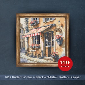 May include: A framed cross-stitch pattern depicting a European cafe scene. The cafe has a red and white striped awning, a small table and chairs, and flower boxes. The text "Cafe" is above the door. The image is labeled "PDF Pattern (Color + Black & White) - Pattern Keeper".