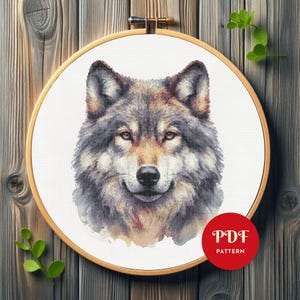 Puede incluir: Un patrón de punto de cruz que presenta un primer plano detallado de la cara de un lobo. El lobo tiene un pelaje gris y marrón con un hocico blanco y una expresión suave y gentil. El patrón está en formato PDF.