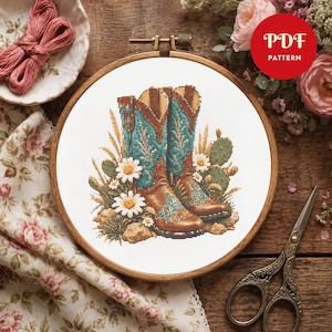 May include: A cross-stitch pattern in a wooden embroidery hoop featuring two brown and turquoise cowboy boots surrounded by white daisies, green cacti, and tall grasses. The design is printed on white fabric with the text PDF PATTERN.