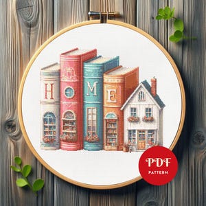 May include: Cross-stitch pattern featuring a stack of colorful books spelling "HOME" and a small house. The design is set within a wooden embroidery hoop, with a red "PDF PATTERN" circle in the corner. The books are in shades of pink, blue, and orange.