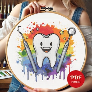 May include: A cross-stitch pattern in a wooden embroidery hoop featuring a smiling white tooth character centered against a vibrant, multi-colored watercolor-style splash background, flanked by a dental pick and a dental mirror tool.