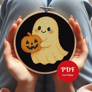 May include: A cross-stitch design featuring a smiling ghost and a pumpkin, set in a black background within a wooden embroidery hoop. The ghost is pale yellow, and the pumpkin is orange. The text "PDF PATTERN" is in a red circle.