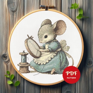 May include: A cross stitch pattern featuring a gray mouse wearing a blue dress and white apron, holding a small embroidery hoop. Beside the mouse is a wooden spool of thread. A red circular badge with the white text PDF PATTERN is included.