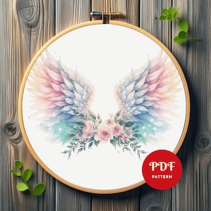 Puede incluir: Un patrón de punto de cruz de alas de ángel en un bastidor de bordado de madera. Las alas están en tonos pastel de rosa, azul y blanco. Debajo de las alas hay flores rosas y hojas verdes. Un círculo rojo con "PDF PATTERN" está en la parte inferior derecha.