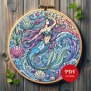 May include: A circular embroidery hoop containing a cross-stitch pattern of a mermaid with flowing pink and blue hair, surrounded by stylized ocean waves, bubbles, and seashells in shades of blue, pink, and purple, with a red PDF PATTERN label.