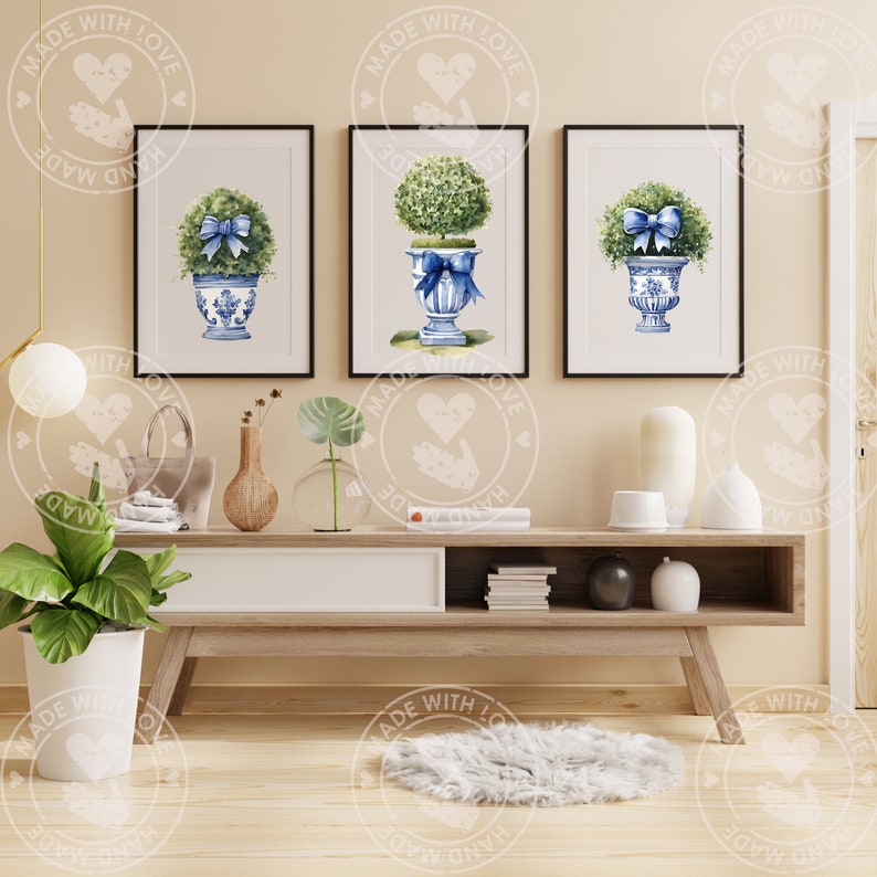 K&ouml;nnte beinhalten: Drei Aquarelle von Topiary-Pflanzen in blau-wei&szlig;en Keramikbeh&auml;ltern mit blauen Schleifen. Die Bilder sind gerahmt und an einer hellbeigen Wand &uuml;ber einem Holz-Konsolentisch aufgeh&auml;ngt.