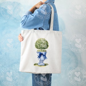 K&ouml;nnte beinhalten: Eine wei&szlig;e Einkaufstasche mit einer blauen und wei&szlig;en Aquarell-Illustration eines Topfbaums mit einer blauen Schleife. Die Tasche hat einen langen Henkel und wird von einer Person gehalten.