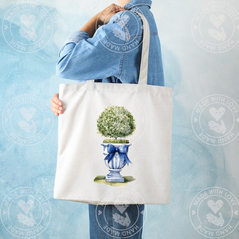 K&ouml;nnte beinhalten: Eine wei&szlig;e Einkaufstasche mit einer blauen und wei&szlig;en Aquarell-Illustration eines Topfbaums mit einer blauen Schleife. Die Tasche hat einen langen Henkel und wird von einer Person gehalten.