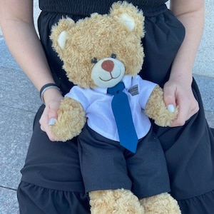 May include: A brown teddy bear wearing a white shirt with a blue tie and black pants. The bear is sitting on a person's lap.