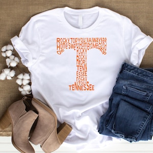 以下が含まれることがあります： 「Rocky Top」「Tennessee」「Home Sweet Home」の文字が入ったオレンジ色の「T」字型のグラフィックが特徴の白いTシャツ。青いジーンズとベージュのアンクルブーツとコーディネートされています。